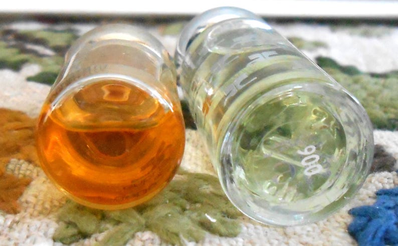 May include: Two clear glass perfume bottles, one filled with amber liquid and the other with a pale green liquid. The bottles are lying on a patterned surface. The bottom of one bottle has the number "906" printed on it.