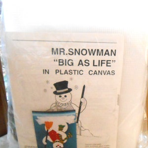 Op de afbeelding: Een plastic canvas knutselset met een sneeuwpop met een zwarte hoge hoed, rode sjaal en bezem. De set bevat acryl garen, een plastic canvas, een patroonkaart en instructies. De set is getiteld "Mr. Snowman "Big as Life" in Plastic Canvas."