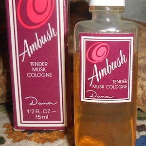 Puede incluir: Un frasco de vidrio transparente de colonia Ambush Tender Musk de Dana con tapa blanca, que contiene líquido ámbar. Al lado hay una caja rectangular a juego en color púrpura oscuro con un logotipo circular rosa, texto blanco y 15 ml.
