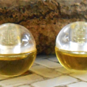 May include: Two clear glass perfume bottles with gold lids and a yellow liquid inside. The bottles are sitting on a tiled surface.