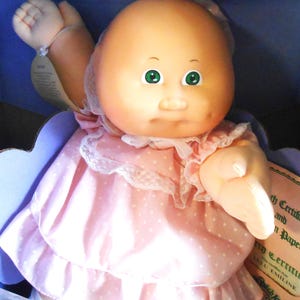 Puede incluir: Una muñeca Cabbage Patch Kids con cabeza calva y ojos verdes. La muñeca lleva un vestido rosa con lunares blancos y ribete de encaje. La muñeca está en una caja azul.