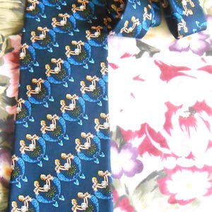 May include: Navy blue necktie with a repeating pattern of mermaids. Each mermaid has blonde hair and a blue tail, holding a seashell. The tie is made of a smooth fabric and is suitable for formal or casual wear.