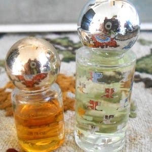 May include: Two small perfume bottles with spherical silver caps. One bottle contains amber liquid and the other contains a clear liquid with a pattern. The amber bottle is labeled "1/8 fl oz".