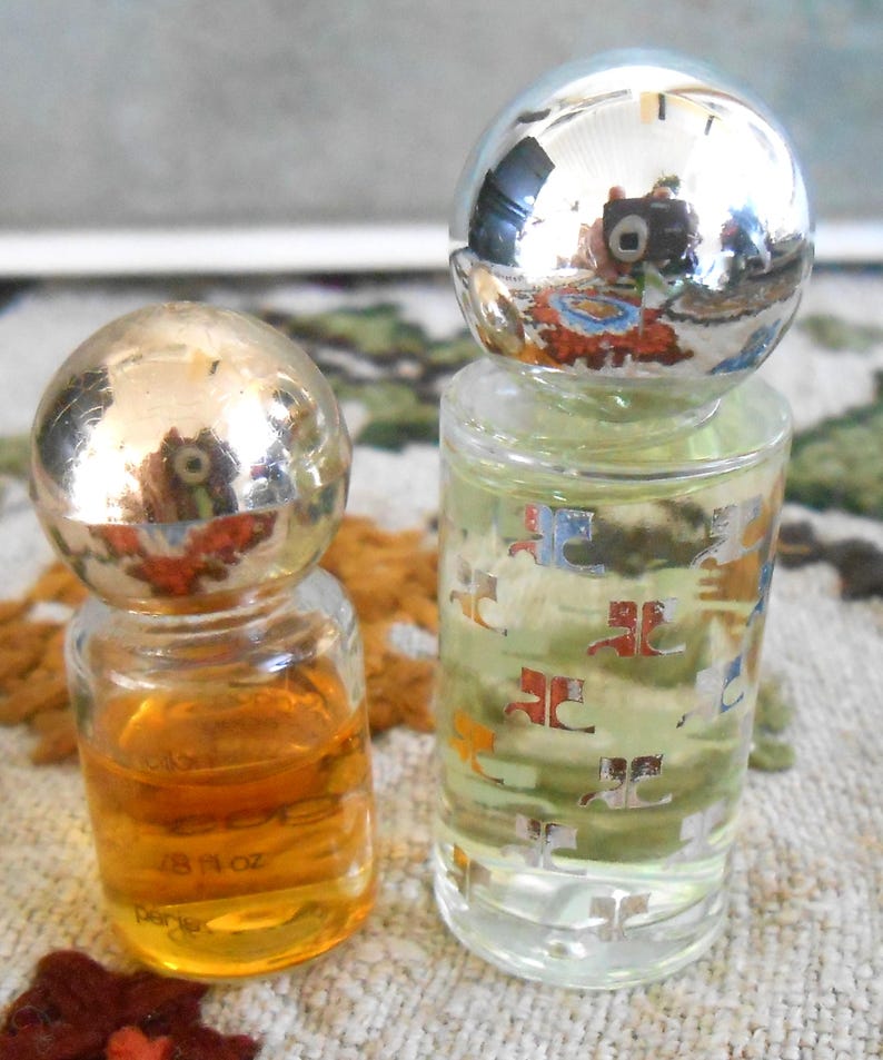May include: Two small perfume bottles with spherical silver caps. One bottle contains amber liquid and the other contains a clear liquid with a pattern. The amber bottle is labeled "1/8 fl oz".