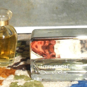 May include: Four miniature perfume bottles with varying designs. The bottles contain amber-colored liquid. One bottle has a black cap, another has a clear, rounded top, and a third has a silver cap with the word "INTRUSION" printed on it. The last bottle has a flower-shaped top.