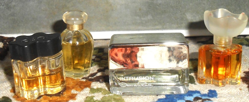 May include: Four miniature perfume bottles with varying designs. The bottles contain amber-colored liquid. One bottle has a black cap, another has a clear, rounded top, and a third has a silver cap with the word "INTRUSION" printed on it. The last bottle has a flower-shaped top.
