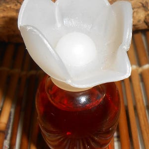 Op de afbeelding: Een klein glazen parfumflesje gevuld met een dieprode vloeistof. Het flesje heeft een doorzichtige, doorschijnende dop in de vorm van een vijfbladige bloem met een afgerond wit midden, rustend op de smalle hals van de ronde glazen houder.