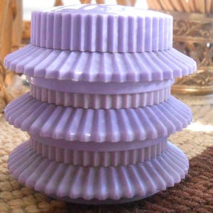 Puede incluir: Una pila de cuatro objetos decorativos escalonados de color lavanda. Cada nivel tiene un diseño acanalado en forma de abanico. El objeto está hecho de un material sólido y liso y está sobre un fondo neutro.