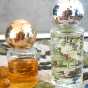 May include: Two small perfume bottles. One has a gold spherical cap and contains amber liquid. The other has a silver spherical cap and a clear bottle with a pattern. Both bottles are on a patterned surface.