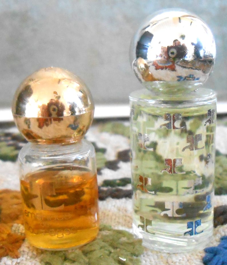 May include: Two small perfume bottles. One has a gold spherical cap and contains amber liquid. The other has a silver spherical cap and a clear bottle with a pattern. Both bottles are on a patterned surface.