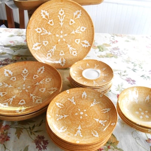 May include: A collection of ceramic dinnerware featuring a golden-yellow textured glaze with a white, stylized floral or snowflake pattern. The set includes large dinner plates, smaller salad plates, saucers with a white center, and deep bowls.
