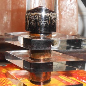 Puede incluir: Un cilindro de vidrio transparente con una parte superior negra y marrón se asienta sobre una pila de tres cuadrados de acrílico transparente. El cilindro está sobre una superficie colorida.