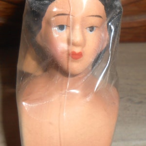 Op de afbeelding: Een vintage poppenhoofd, verpakt in doorzichtig plastic. De pop heeft donker haar, blauwe ogen en rode lippen. De huidskleur is licht perzik. De buste van de pop is zichtbaar en de achtergrond is onscherp.