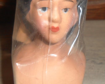 Może przedstawiać: Głowa vintage lalki, zapakowana w przezroczysty plastik. Lalka ma ciemne włosy, niebieskie oczy i czerwone usta. Odcień skóry jest jasny brzoskwiniowy. Widoczny jest biust lalki, a tło jest rozmyte.