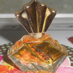 May include: A small, faceted perfume bottle with a gold-colored, fan-shaped cap. The bottle contains a golden liquid and has the word "Violet" in black script. The brand name "ANNA MARCHETTI" is printed on the bottle.
