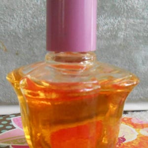 May include: A small, clear glass perfume bottle with a pink cap. The bottle is filled with a golden-orange liquid. The bottle has a unique, flared shape. The background is a floral pattern.