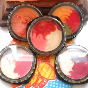 Puede incluir: Cinco pequeños recipientes redondos con tapas transparentes, cada uno contiene un remolino de bálsamo labial de diferentes colores. Los colores incluyen rojo, rosa, blanco, naranja y amarillo.