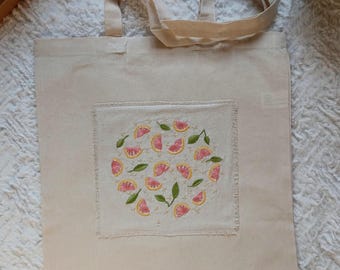 May include: Cream-colored tote bag with embroidered citrus fruit design. The embroidery features pink grapefruit slices and green leaves arranged in a circular pattern. The bag has two handles and a rectangular patch with the embroidery.