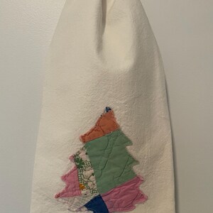 May include: A white hand towel featuring a patchwork Christmas tree design. The tree is made from various fabric scraps in shades of pink, green, blue, and orange. The towel hangs from a silver ring.
