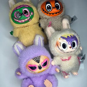May include: Four plush toys in the shape of rabbits, each with a unique monster-themed face. The toys are in yellow, gray, purple, and white, with details like a pumpkin, a brain, and a spiderweb. The toys have bunny ears and small feet.