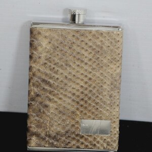 May include: A rectangular flask with a silver cap and trim. The body of the flask is covered in a textured, tan material with a scale-like pattern. A small, rectangular silver plate is affixed to the front.