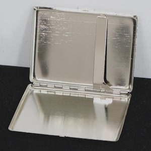 May include: Open, silver-colored metal business card case. The case is rectangular with a hinged lid and a small interior divider. The interior has a shiny, reflective surface. The case is open, revealing the inside.
