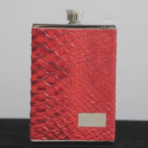 May include: A rectangular flask with a red, textured exterior, possibly faux leather, and a silver-colored cap and trim. The flask has a small, rectangular silver-colored label near the bottom. The flask is designed to hold liquids.