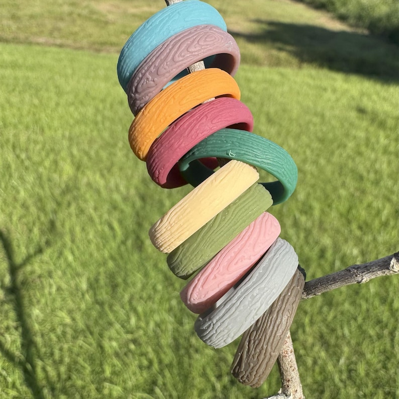 May include: A collection of colorful, textured bracelets in various shades, including blue, purple, orange, and green. The bracelets have a wood-grain pattern and are displayed on a small branch against a blurred green background.