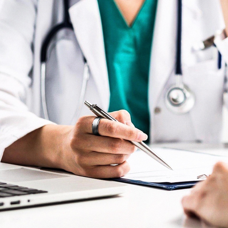 May include: A doctor wearing a white coat and a teal scrub top is sitting at a desk with a laptop and a clipboard. The doctor is holding a silver pen and is pointing at the clipboard.