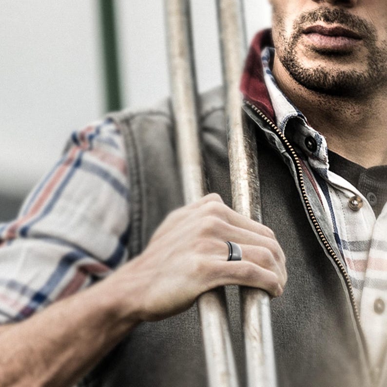 May include: A close-up of a man's hand wearing a dark gray silicone ring. The ring has a beveled edge and a polished finish.