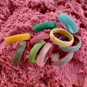 May include: A collection of colorful, textured bangles in shades of yellow, green, pink, blue, and gray. The bangles have a wood grain pattern and are arranged on a bed of pink sand.