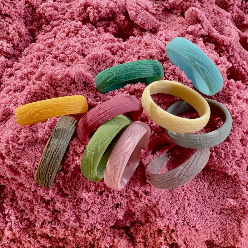May include: A collection of colorful, textured bangles in shades of yellow, green, pink, blue, and gray. The bangles have a wood grain pattern and are arranged on a bed of pink sand.