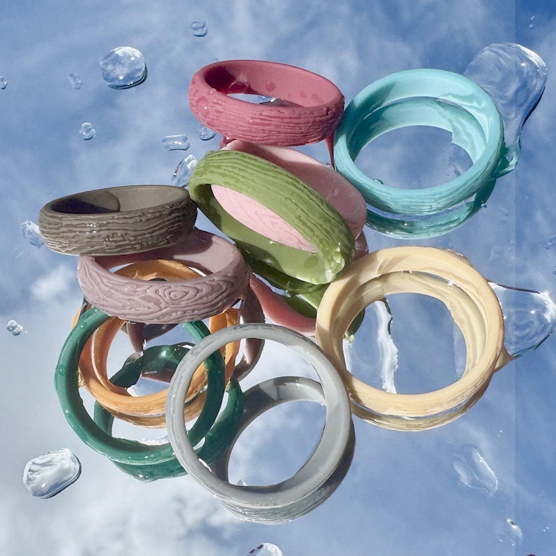 May include: A collection of colorful rings in various designs and textures. The rings are in shades of pink, green, blue, yellow, and gray. Some rings have a wood-grain texture, while others are smooth. The rings are arranged on a reflective surface with water droplets.