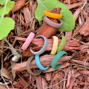 May include: A collection of colorful silicone rings in various shades, including yellow, pink, gray, blue, green, and brown. The rings have a textured wood grain pattern and are arranged on a bed of wood chips.