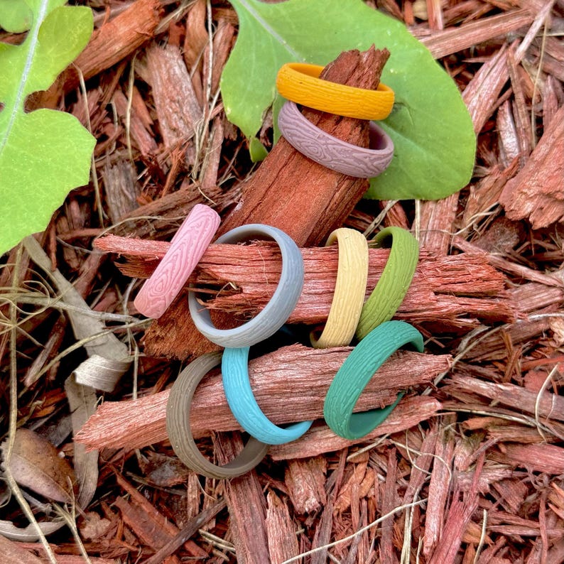 May include: A collection of colorful silicone rings in various shades, including yellow, pink, gray, blue, green, and brown. The rings have a textured wood grain pattern and are arranged on a bed of wood chips.