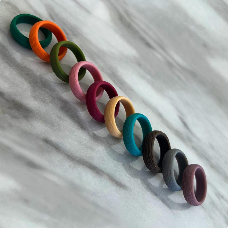 May include: A collection of colorful silicone rings in various shades, including green, orange, pink, and blue. The rings have a textured, wood-like appearance. The rings are arranged diagonally on a marble surface.