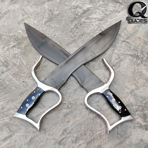 May include: Two crossed black bladed knives with silver handles and black grips. The handles have a curved design and are decorated with small, circular silver accents. The blades are wide and have a dark finish. The logo "Q Blades" is in the upper right corner.