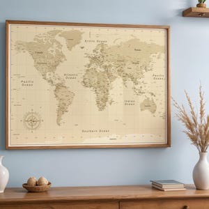 Op de afbeelding: Een wereldkaart met lijst in een warme beige tint, met gedetailleerde geografische kenmerken en landnamen. De kaart is ingelijst in een houten frame en hangt aan een lichtblauwe muur. Een kompasroos is te zien in de linker benedenhoek. Decoratieve items staan op een houten oppervlak eronder.