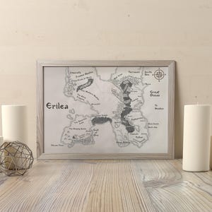 May include: A framed map of Erilea, a fantasy world, in grayscale. The map is detailed with geographical features and text labels. The frame is light-coloured. Several white pillar candles and a geometric metal sphere are in front of the map.