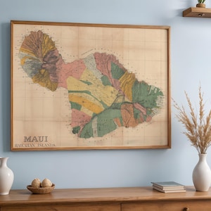 Puede incluir: Un mapa vintage enmarcado de Maui, Islas Hawaianas, con un marco de madera. El mapa presenta una ilustración detallada y colorida de la topografía de la isla. El mapa se muestra en una pared azul claro, con un jarrón blanco y otros elementos decorativos.