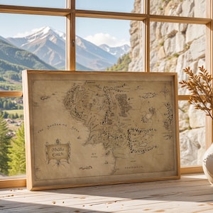 May include: A framed map of Middle Earth, displayed on a wooden surface near a window. The map is in sepia tones with intricate details, including text and illustrations. Two white vases with dried plants are on the right. The background shows a mountain range.