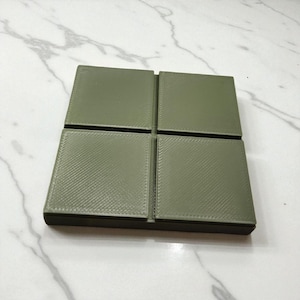 Op de afbeelding: Een vierkant, olijfgroen object met vier even grote, getextureerde vierkanten. De vierkanten zijn in een rasterpatroon gerangschikt, met een donkere rand. Het kan een onderzetter of een vergelijkbaar item zijn.