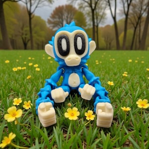 May include: A blue articulated monkey figurine with white accents, sitting in a grassy field with yellow flowers. The monkey has large black eyes and a white belly button. The background features trees and a blurred green landscape.