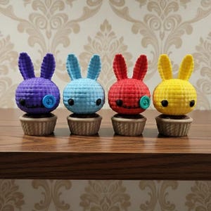 May include: Four crochet bunny figurines in purple, blue, red, and yellow, each with button eyes and a button nose. They sit on small, brown cupcake-like bases, arranged on a wooden surface against a patterned wallpaper background.