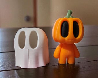 May include: Two Halloween-themed figurines: a white ghost and an orange pumpkin-headed character. The ghost is a simple, rounded shape with two eye holes. The pumpkin figure has black eyes and a green stem.