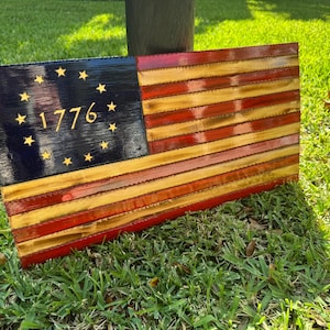 May include: Wooden American flag with a distressed finish. The flag features a blue canton with gold stars and the year "1776". The stripes are red and yellow, creating a rustic, patriotic decor item.