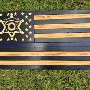 May include: Wooden American flag with a black background, natural wood stripes, and a blue line. The flag features a sheriff's badge in the star field. The stars are cut out of the black wood, and the stripes are made of natural wood.