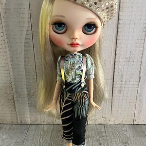 May include: A doll with blonde hair wearing a gold sequin beret, a blue and white floral print shirt with gold suspenders, black and white striped pants, and gold shoes.