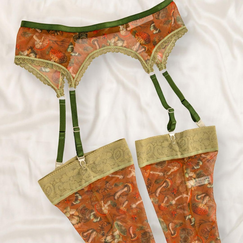 May include: A garter belt and matching thigh-high stockings set featuring a sheer orange fabric with a detailed mushroom and botanical print. The set includes dark green elastic straps, adjustable metal sliders, and wide lace trim edges.