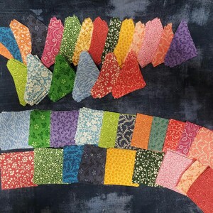 May include: A collection of colorful fabric squares arranged on a dark blue surface. The squares feature various floral and patterned designs in shades of blue, green, orange, red, and purple. These fabric pieces are ideal for quilting and crafting projects.
